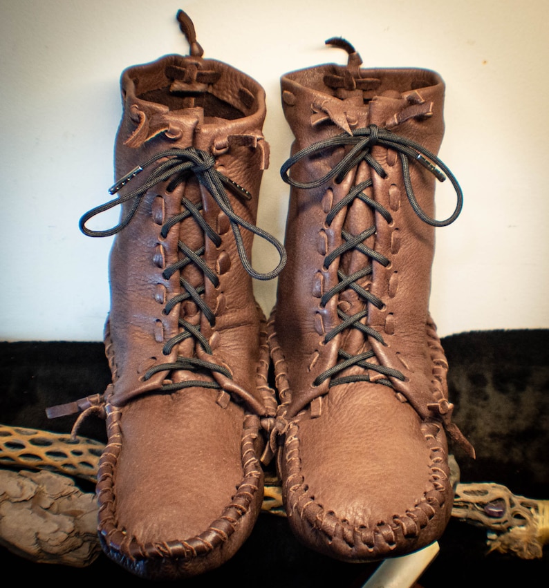 Puede incluir: Un par de mocasines de cuero marr&oacute;n con cordones negros. Los mocasines tienen una costura cosida alrededor del borde de la suela y est&aacute;n atados en el tobillo.