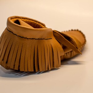 May include: A single tan leather moccasin with a fringed vamp and a soft, rounded toe. The shoe has a simple design with visible stitching and a leather tie. The moccasin is likely for a baby or toddler.