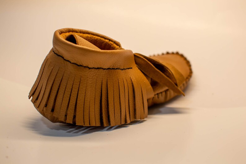 May include: A single tan leather moccasin with a fringed vamp and a soft, rounded toe. The shoe has a simple design with visible stitching and a leather tie. The moccasin is likely for a baby or toddler.