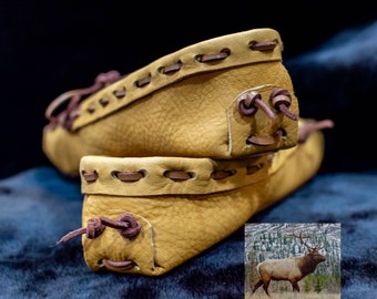 May include: Two pairs of tan leather moccasins with brown stitching and laces. The moccasins are stacked on top of each other. A photo of an elk in a snowy forest is in the lower right corner of the image.
