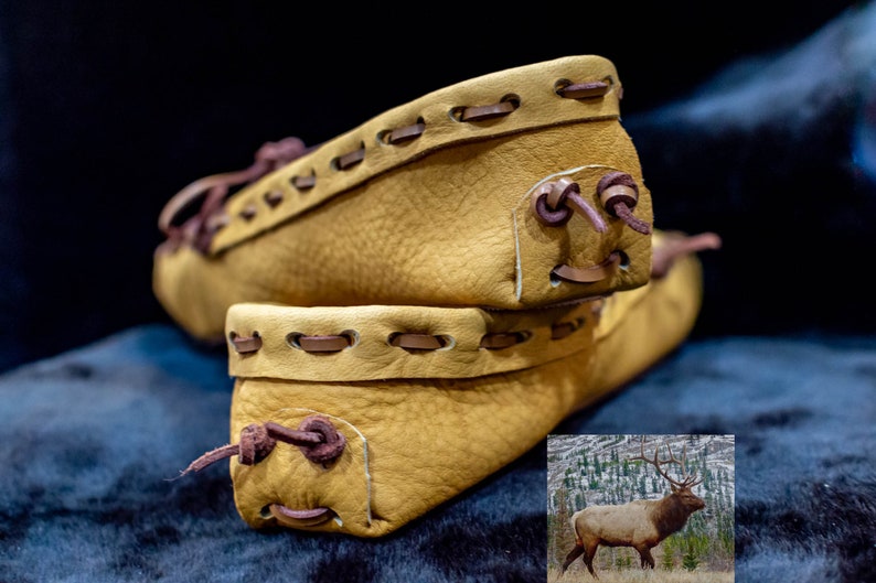 May include: Two pairs of tan leather moccasins with brown stitching and laces. The moccasins are stacked on top of each other. A photo of an elk in a snowy forest is in the lower right corner of the image.
