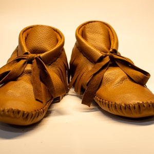 May include: A pair of tan leather baby moccasins. The shoes have a soft, textured appearance with a fringed collar and a ribbon tie. The soles are stitched with a visible seam. These baby shoes are designed for comfort and style.
