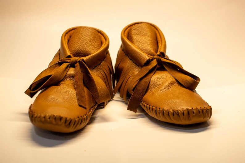 May include: A pair of tan leather baby moccasins. The shoes have a soft, textured appearance with a fringed collar and a ribbon tie. The soles are stitched with a visible seam. These baby shoes are designed for comfort and style.