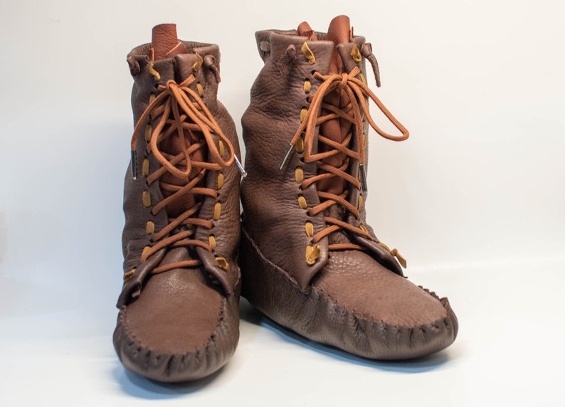 Puede incluir: Un par de mocasines de cuero marr&oacute;n con cordones. Los mocasines tienen un eje alto y est&aacute;n atados con cordones marrones.