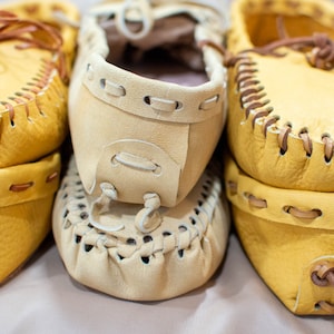 May include: Three pairs of yellow leather moccasins with brown stitching and laces. The moccasins are stacked on top of each other.