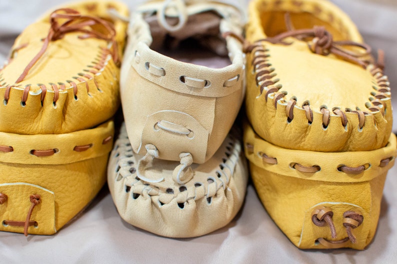 May include: Three pairs of yellow leather moccasins with brown stitching and laces. The moccasins are stacked on top of each other.