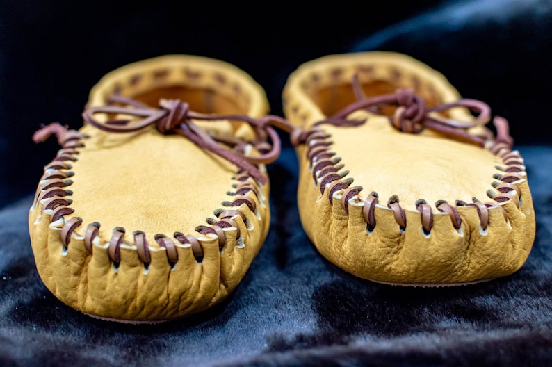 May include: A pair of tan leather moccasins with brown leather stitching and laces.