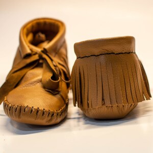 May include: A pair of soft, brown leather baby moccasins. One shoe is laced up, while the other is a fringed ankle cuff. The shoes are made of a smooth, light brown leather and have visible stitching.