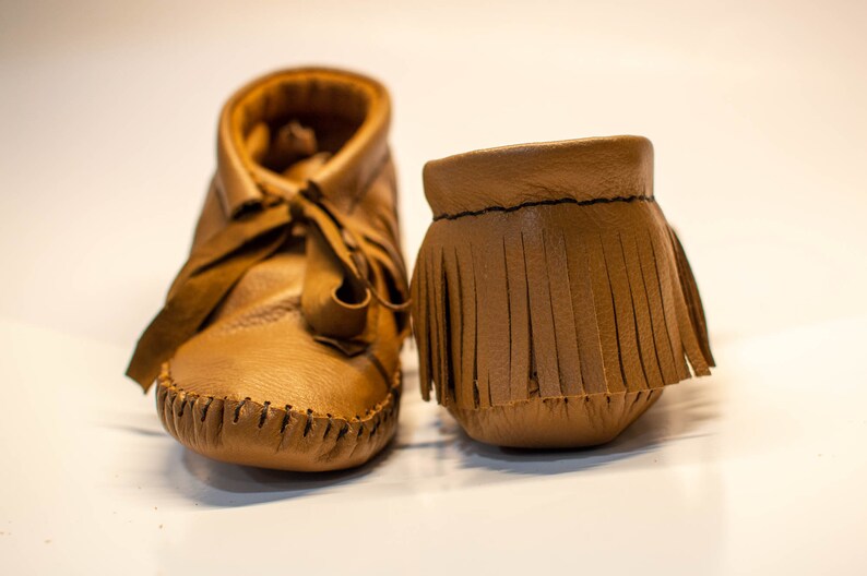 May include: A pair of soft, brown leather baby moccasins. One shoe is laced up, while the other is a fringed ankle cuff. The shoes are made of a smooth, light brown leather and have visible stitching.