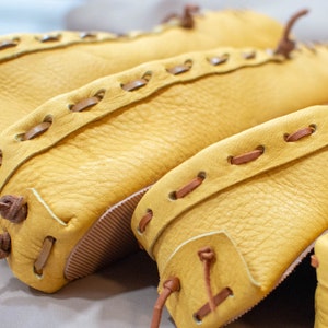 May include: Close-up of three pairs of yellow leather moccasins with brown leather stitching. The moccasins are stacked on top of each other.