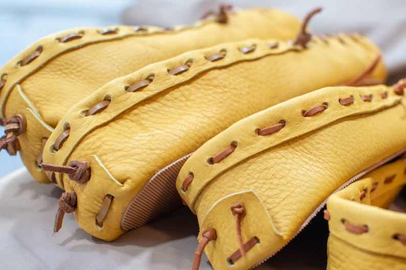 May include: Close-up of three pairs of yellow leather moccasins with brown leather stitching. The moccasins are stacked on top of each other.