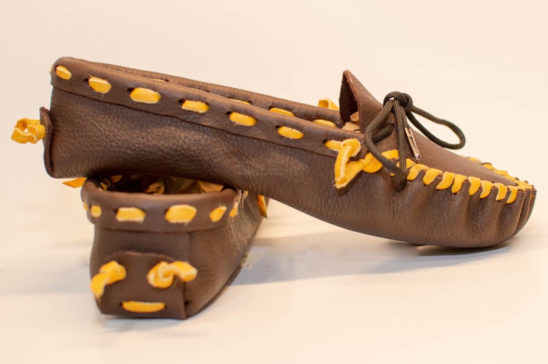 May include: A pair of brown leather moccasins with yellow stitching. The moccasins have a simple design and are tied with a black lace.