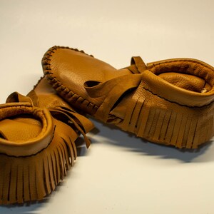 May include: A pair of tan leather moccasins with fringe detailing and a soft, rounded collar. The shoes are made of a smooth, light brown leather and feature a tie closure. The moccasins are designed for infants or toddlers.