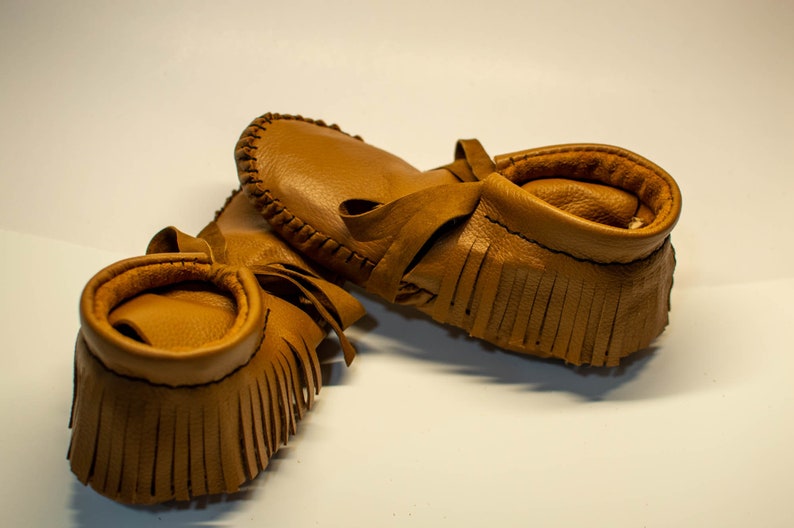May include: A pair of tan leather moccasins with fringe detailing and a soft, rounded collar. The shoes are made of a smooth, light brown leather and feature a tie closure. The moccasins are designed for infants or toddlers.