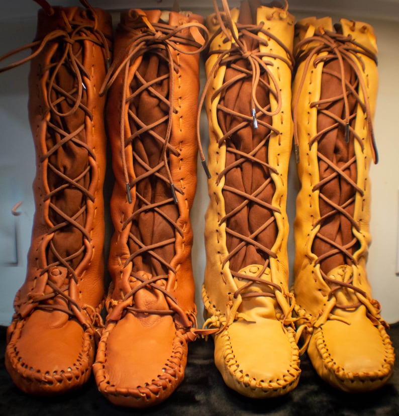 May include: Three pairs of brown leather moccasins with laces. The moccasins are knee-high and have a decorative stitch pattern around the sole.
