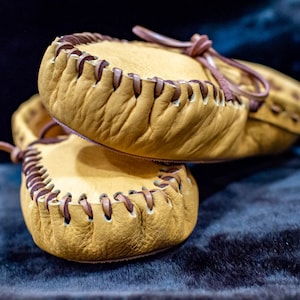 May include: A pair of tan leather moccasins with brown stitching. The moccasins are stacked on top of each other.