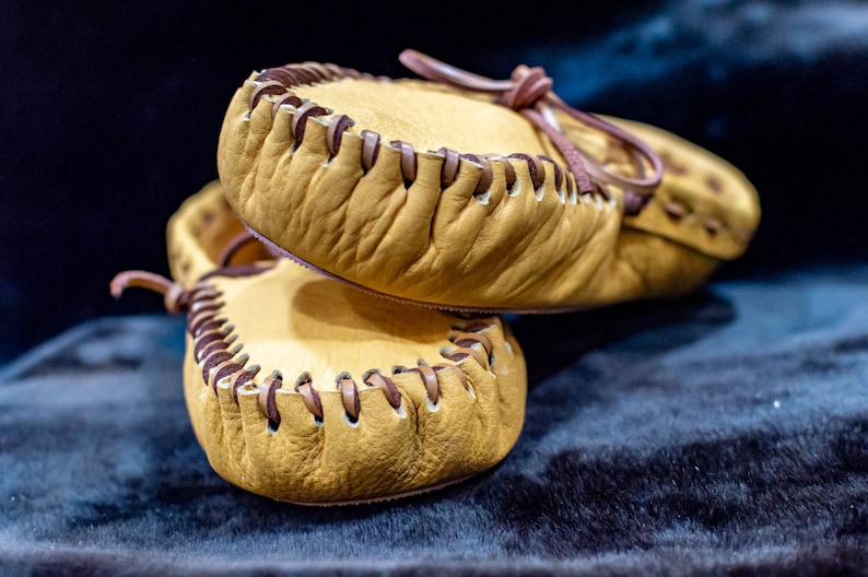 May include: A pair of tan leather moccasins with brown stitching. The moccasins are stacked on top of each other.