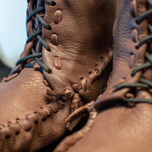 Puede incluir: Primer plano de un par de mocasines de cuero marr&oacute;n con cordones negros. Los mocasines tienen un patr&oacute;n de costura &uacute;nico y est&aacute;n hechos de cuero de alta calidad.