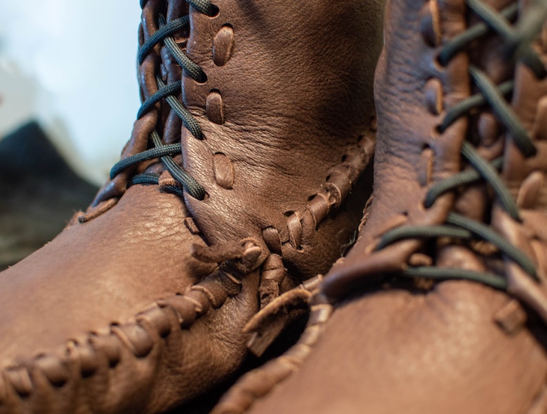 Puede incluir: Primer plano de un par de mocasines de cuero marr&oacute;n con cordones negros. Los mocasines tienen un patr&oacute;n de costura &uacute;nico y est&aacute;n hechos de cuero de alta calidad.