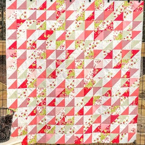 May include: A handmade quilt with a repeating pattern of triangles in red, pink, green, and white floral and patterned fabrics. The quilt is hanging on a black metal rod with black clips, displayed outdoors.