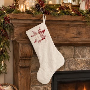 May include: A cream-colored fabric Christmas stocking featuring a red and white plaid star appliqué centered on the upper portion. The stocking includes a simple fabric loop at the top for hanging and has a soft, textured surface appearance.