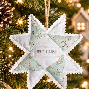 May include: A quilted star-shaped Christmas ornament featuring alternating panels of solid white fabric and light green fabric with a pink snowflake pattern. The center includes a white rectangular patch with the text MERRY CHRISTMAS!