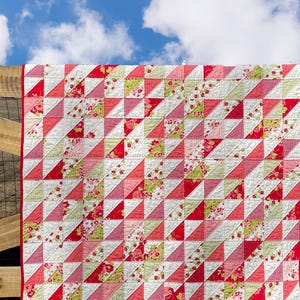 May include: A handmade quilt with a repeating pattern of red, pink, and white triangles, some with floral prints. The quilt has a red border and is displayed outdoors against a blue sky. The logo for Calico Bliss Designs is visible.
