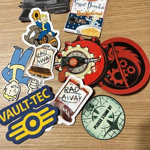 May include: A collection of Fallout themed stickers featuring a Vault-Tec logo, a Pip-Boy, a Nuka-Cola bottle, a Rad-Away bottle, a character riding a deathclaw, a gun, and a "Please Stand By" sign.