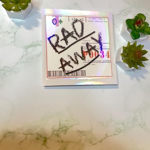 Könnte beinhalten: Ein weißer Aufkleber mit holografischem Finish. Der Aufkleber hat schwarzen Text, der "RAD AWAY" und "P0034" lautet. Der Aufkleber hat auch ein rotes "O+" Symbol und den Text "100 ML".