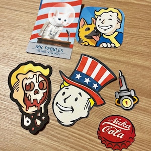 May include: A set of six Fallout themed iron-on patches. The patches feature a cartoon character with a skull, a cartoon character wearing a red, white, and blue top hat, a cartoon character with a dog, a cartoon cat in a space suit, a cartoon rocket ship, and a red and white bottle cap with the text "Nuka Cola".