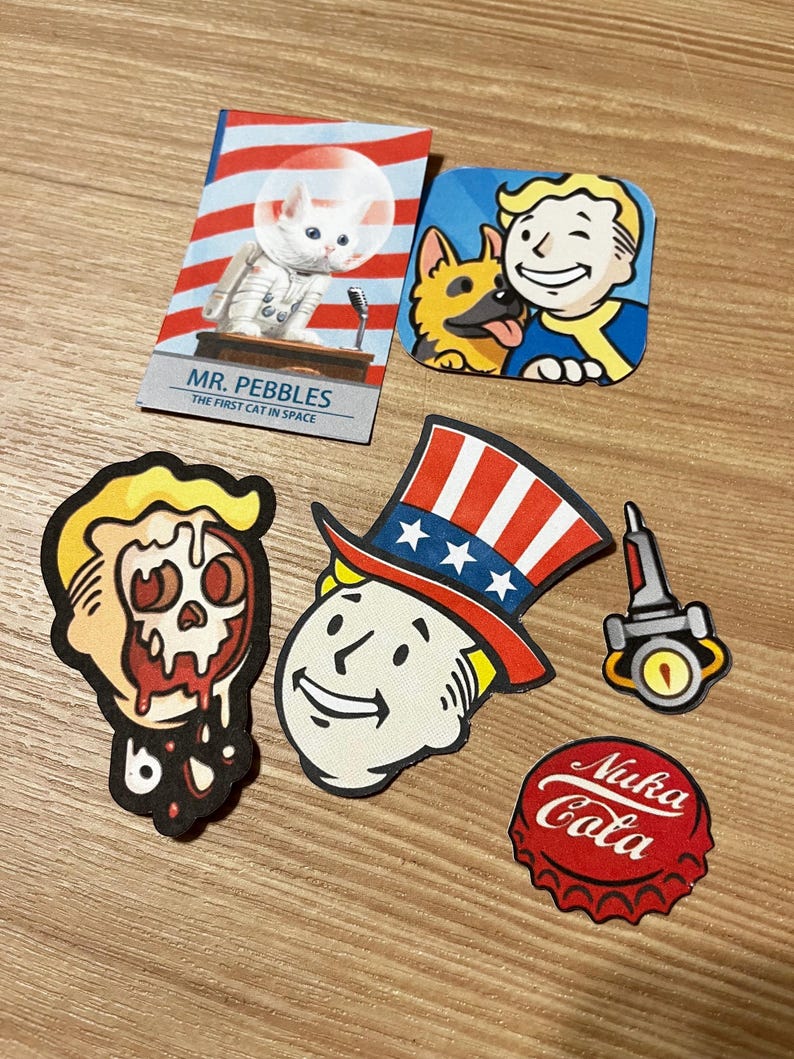May include: A set of six Fallout themed iron-on patches. The patches feature a cartoon character with a skull, a cartoon character wearing a red, white, and blue top hat, a cartoon character with a dog, a cartoon cat in a space suit, a cartoon rocket ship, and a red and white bottle cap with the text "Nuka Cola".