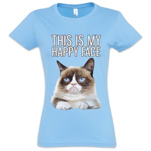 Könnte beinhalten: Hellblaues T-Shirt mit dem Text "THIS IS MY HAPPY FACE" und einer Grafik einer grummeligen Katze. Die Katze hat ein braun-weißes Gesicht mit blauen Augen. Der Text ist in weißen Blockbuchstaben.
