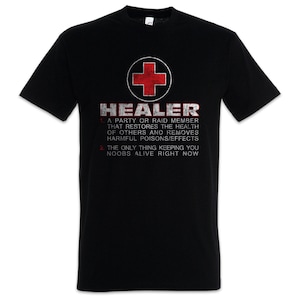 Peut inclure: T-shirt noir avec le mot "HEALER" en texte blanc vieilli et un emblème de croix rouge. Le t-shirt comprend du texte définissant le rôle d'un guérisseur dans un jeu et la phrase "The only thing keeping you noobs alive right now."