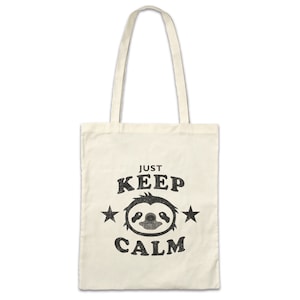 Peut inclure: Sac fourre-tout crème avec texte noir et un motif de paresseux. Le texte "JUST KEEP CALM" est au-dessus et en dessous du visage du paresseux, avec des étoiles de chaque côté. Le sac a deux longues anses.