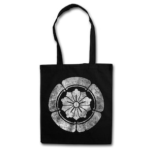 May include: Black tote bag with a distressed white floral crest design. The bag has two black handles and is made of a durable fabric. The design is centered on the front of the bag.