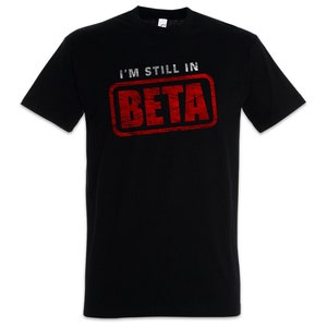 May include: Black crew neck t-shirt featuring the text "I'M STILL IN BETA" in white and red. The text is within a red, distressed-effect rectangle. The shirt is short-sleeved and suitable for casual wear.