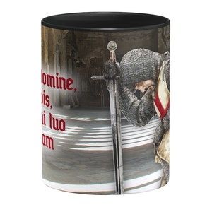 May include: A black ceramic mug with a white interior. The mug features a graphic of a knight in armor kneeling with a sword. The knight is wearing a red and white cross on his chest. The text "Domine, bis, ni tuo am" is printed in red on the mug.