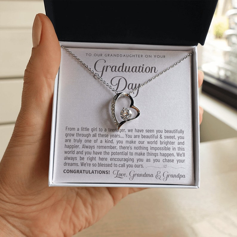 Peut inclure: Un collier pendentif en forme de c&oelig;ur argent&eacute; avec une pierre pr&eacute;cieuse transparente au centre, pr&eacute;sent&eacute; dans un coffret cadeau blanc. Le coffret contient un message pour une petite-fille pour son jour de remise de dipl&ocirc;me. Le texte dit "Graduation Day".