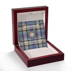 Pode incluir: Um colar dourado com um pingente de pedra transparente, apresentado numa caixa de madeira vermelha escura. A caixa cont&eacute;m um cart&atilde;o com o texto "To My Sassenach" e uma cita&ccedil;&atilde;o sobre o amor. O fundo apresenta um padr&atilde;o xadrez azul e verde.