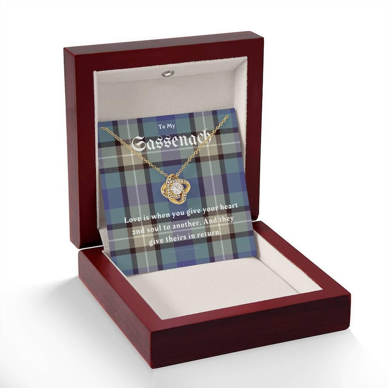 Pode incluir: Um colar dourado com um pingente de pedra transparente, apresentado numa caixa de madeira vermelha escura. A caixa cont&eacute;m um cart&atilde;o com o texto "To My Sassenach" e uma cita&ccedil;&atilde;o sobre o amor. O fundo apresenta um padr&atilde;o xadrez azul e verde.