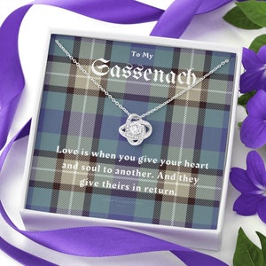 Pode incluir: Um colar de prata com um pendente de n&oacute;, apresentado numa caixa branca com um design xadrez. A caixa diz "To My Sassenach" e inclui a cita&ccedil;&atilde;o "Amar &eacute; quando voc&ecirc; entrega seu cora&ccedil;&atilde;o e alma a outro."