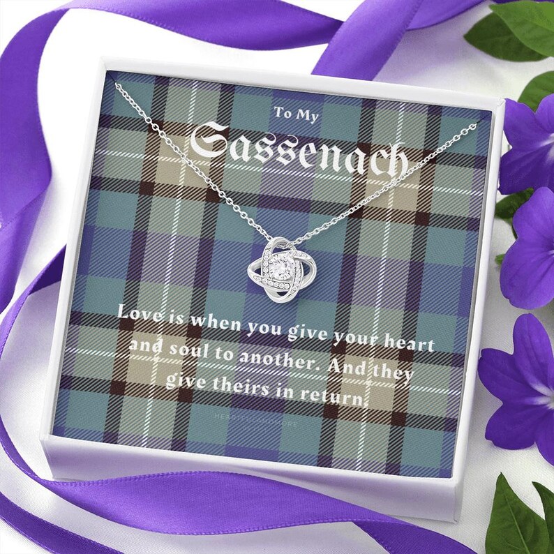 Pode incluir: Um colar de prata com um pendente de n&oacute;, apresentado numa caixa branca com um design xadrez. A caixa diz "To My Sassenach" e inclui a cita&ccedil;&atilde;o "Amar &eacute; quando voc&ecirc; entrega seu cora&ccedil;&atilde;o e alma a outro."