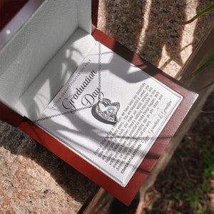 Peut inclure: Un collier pendentif argent&eacute; en forme de c&oelig;ur avec une pierre centrale transparente, pr&eacute;sent&eacute; dans un coffret cadeau rouge. La bo&icirc;te contient une carte avec le texte "Graduation Day" et un message de Grand-m&egrave;re et Grand-p&egrave;re.