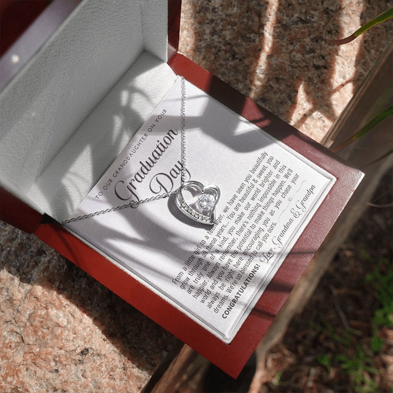 Peut inclure: Un collier pendentif argent&eacute; en forme de c&oelig;ur avec une pierre centrale transparente, pr&eacute;sent&eacute; dans un coffret cadeau rouge. La bo&icirc;te contient une carte avec le texte "Graduation Day" et un message de Grand-m&egrave;re et Grand-p&egrave;re.