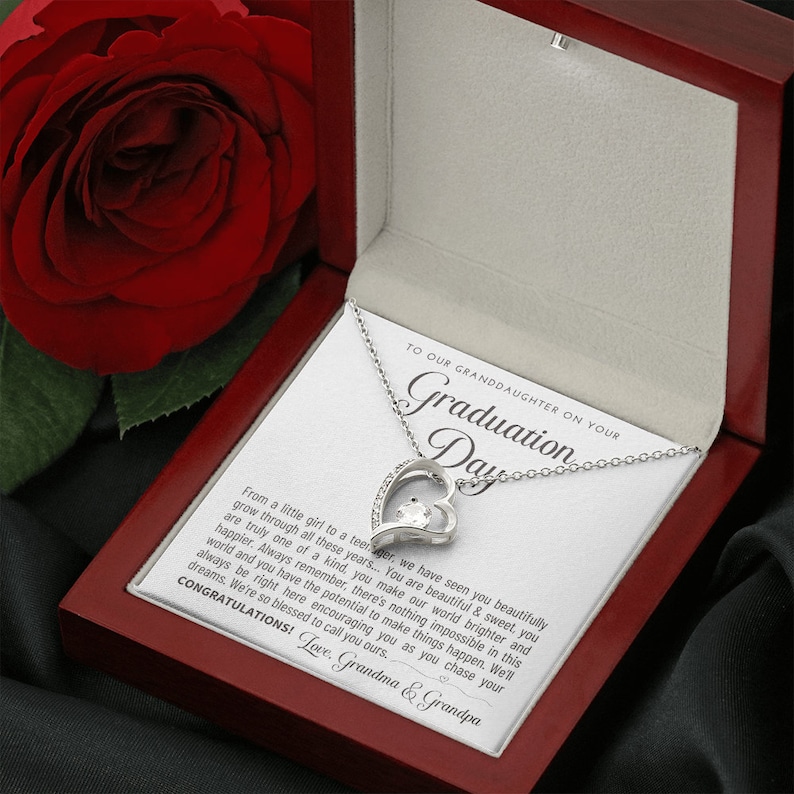 Peut inclure: Un collier pendentif argent&eacute; en forme de c&oelig;ur avec une pierre pr&eacute;cieuse transparente, pr&eacute;sent&eacute; dans un coffret cadeau en bois rouge ouvert. Le coffret contient une carte avec le texte "TO OUR GRANDDAUGHTER ON YOUR Graduation Day."