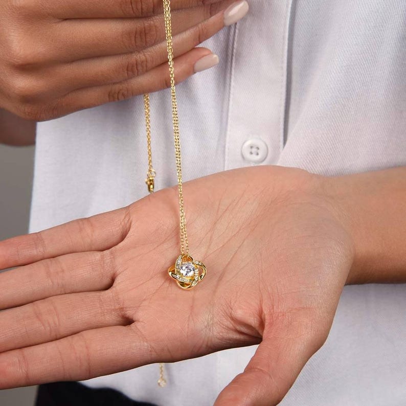 Pode incluir: Um colar dourado com um pingente em forma de n&oacute;, com uma pedra preciosa transparente central e pedras transparentes menores. O colar &eacute; segurado em uma m&atilde;o aberta, contra uma camisa branca.