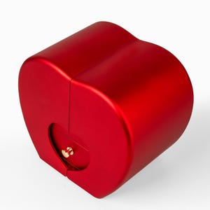 Pode incluir: Uma caixa vermelha em forma de cora&ccedil;&atilde;o com acabamento met&aacute;lico. A caixa tem uma pequena abertura redonda com um fecho dourado. A caixa &eacute; projetada com uma forma curva e cil&iacute;ndrica.