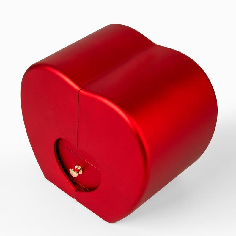 Pode incluir: Uma caixa vermelha em forma de cora&ccedil;&atilde;o com acabamento met&aacute;lico. A caixa tem uma pequena abertura redonda com um fecho dourado. A caixa &eacute; projetada com uma forma curva e cil&iacute;ndrica.