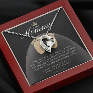 May include: A silver necklace with a heart pendant and a small clear stone in the center. The necklace is in a red box with a message that reads "To My Mommy I may just be a bump, but I love you more than you'll ever know. Please keep this necklace close to your heart until I can meet you and rest in your arms. Love, kisses & kicks from your tummy. Joolnco.com"