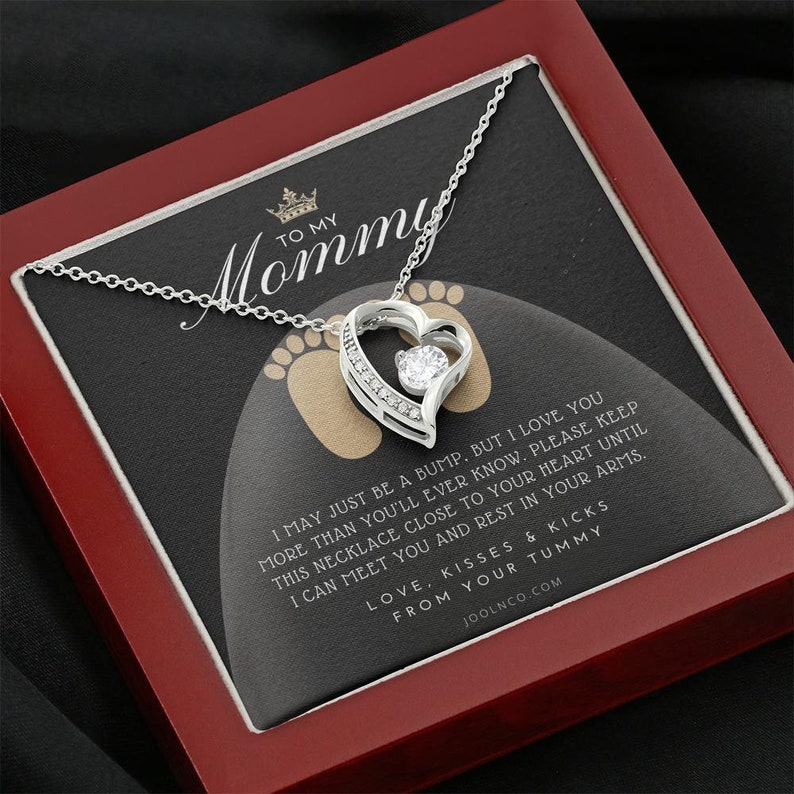 May include: A silver necklace with a heart pendant and a small clear stone in the center. The necklace is in a red box with a message that reads "To My Mommy I may just be a bump, but I love you more than you'll ever know. Please keep this necklace close to your heart until I can meet you and rest in your arms. Love, kisses & kicks from your tummy. Joolnco.com"